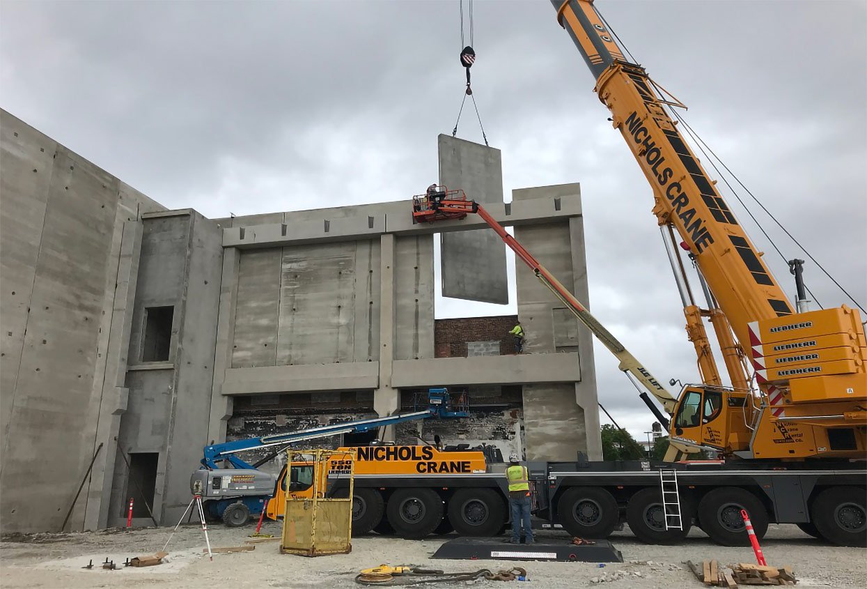 A yellow crane lifting a large concrete wall panel into place on a construction site.