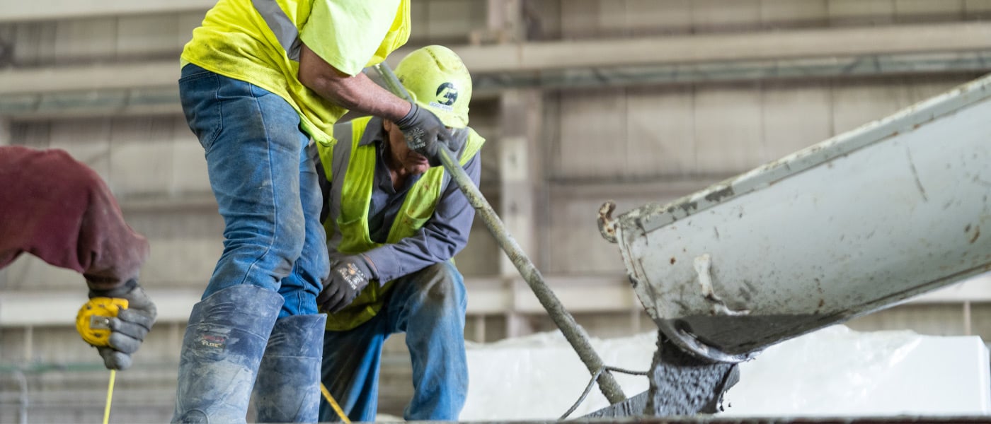 Two men wearing high-viz vests, hard hats, and blue jeans work with wet concrete in a warehouse.